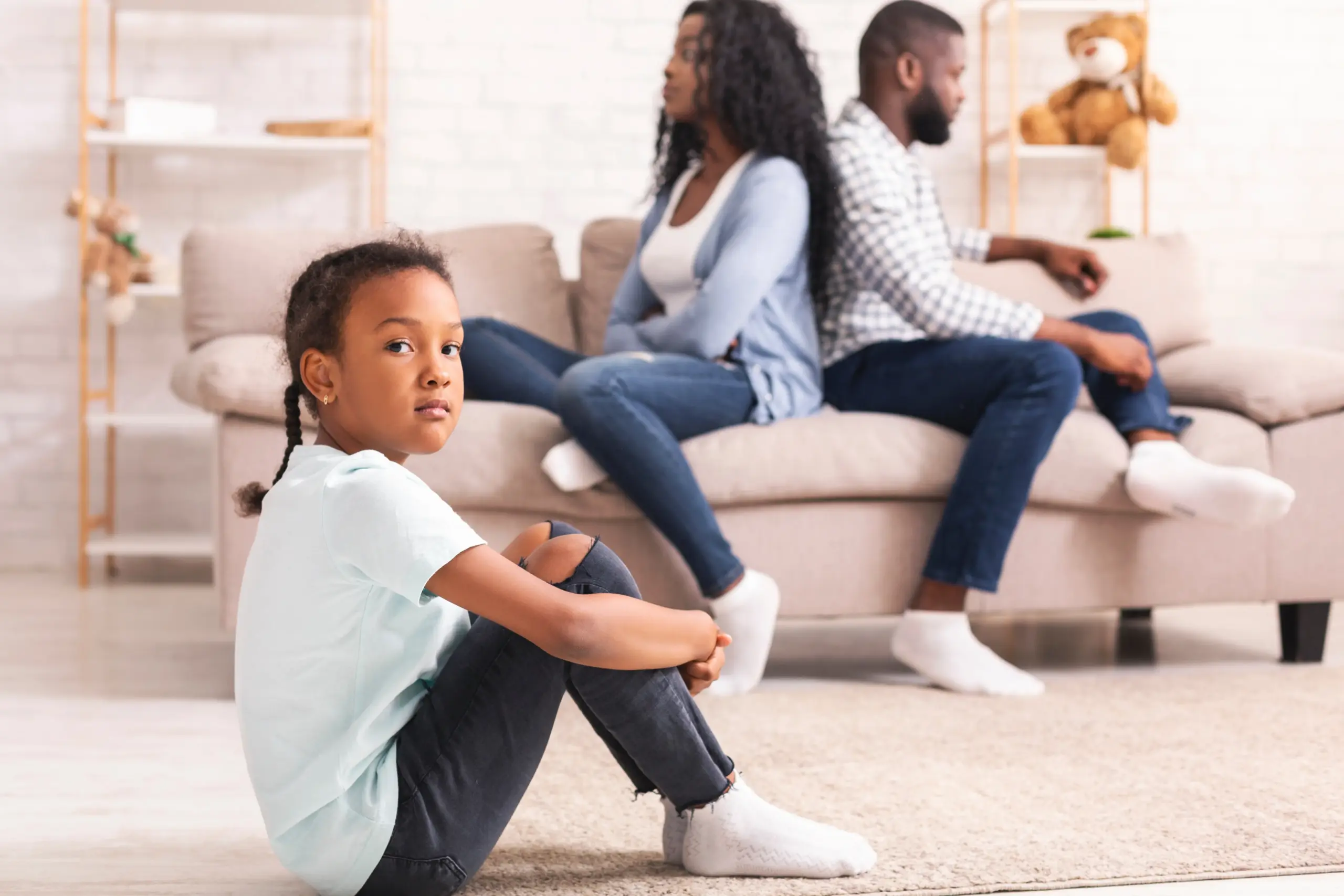 Girl looking at camera with her knees to her chest as arguing parents sit on a couch back to back, not talking.